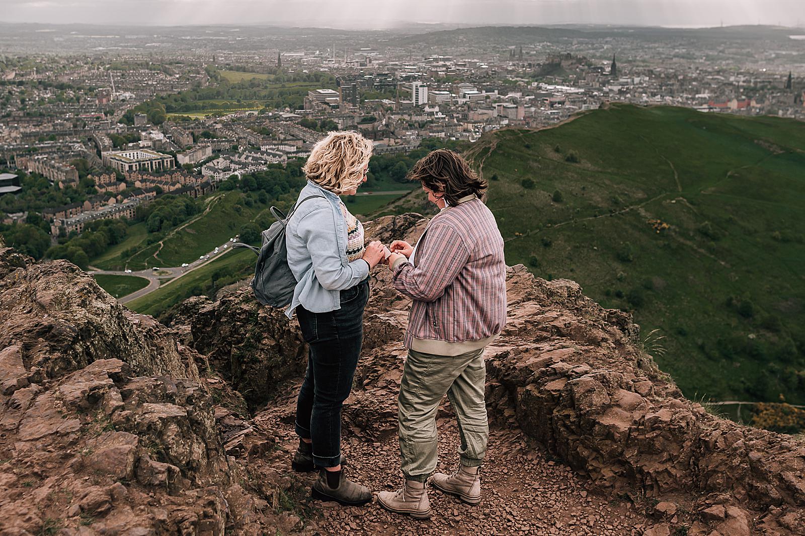 couple standing on rocky hillside Arthurs seat for surprise proposal photo shoot by Edinburgh proposal photographer on a windy moody day in scotland