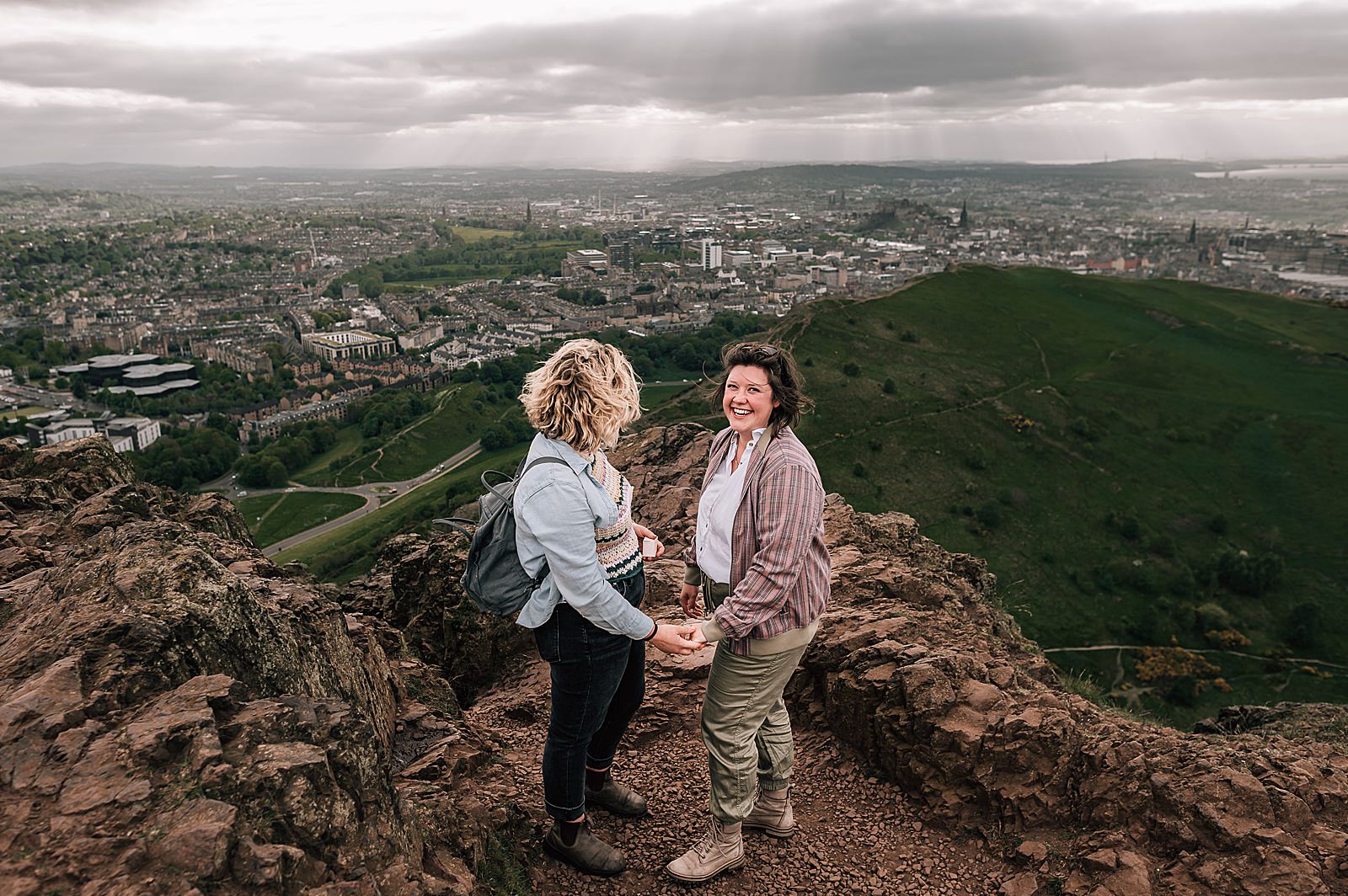 couple holding hands while standing on top of Arthurs seat Edinburgh on a windy day during their surprise proposal photo shoot Edinburgh