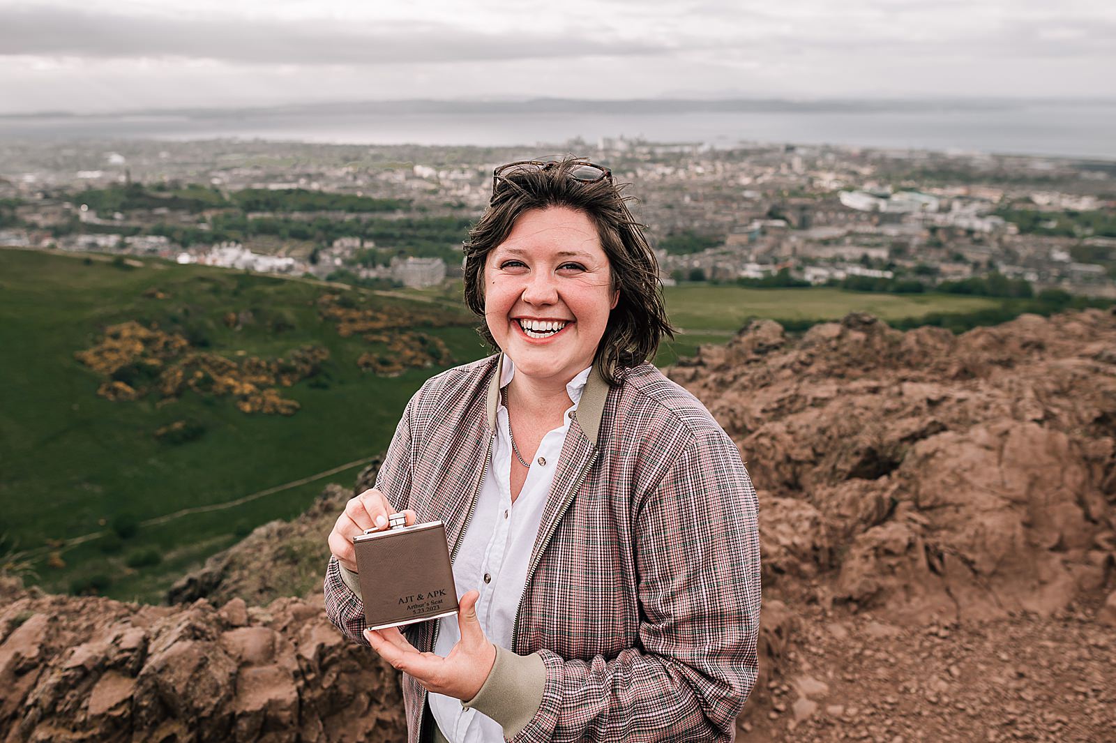 girl holding personalised hip flask and smiling while standing on top of Arthurs seat Edinburgh during surprise marriage proposal Edinburgh captured by Scottish proposal photographer