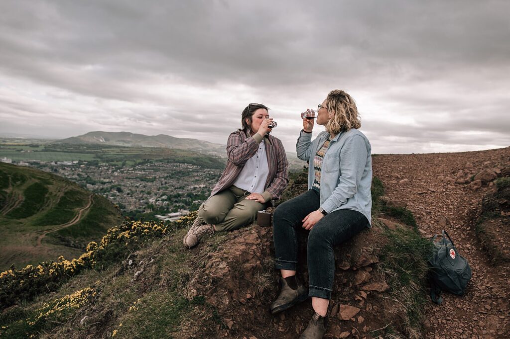 couple sipping a dram of whisky on top of Arthurs seat during their proposal photo shoot Edinburgh taken by proposal photographer scotland