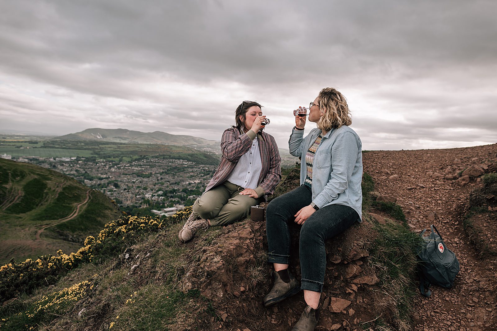 couple sipping a dram of whisky on top of Arthurs seat during their proposal photo shoot Edinburgh taken by proposal photographer scotland