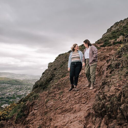 lesbian couple holding hands walking down a steep rocky path on Arthurs seat Edinburgh after engagement photo shoot