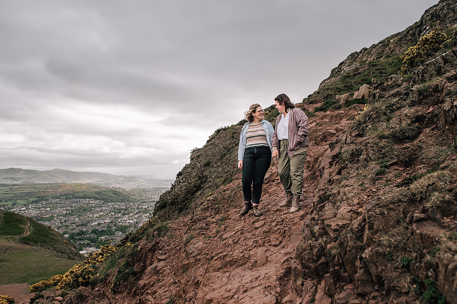 couple holding hands walking down a steep rocky path on Arthurs seat Edinburgh after engagement photo shoot