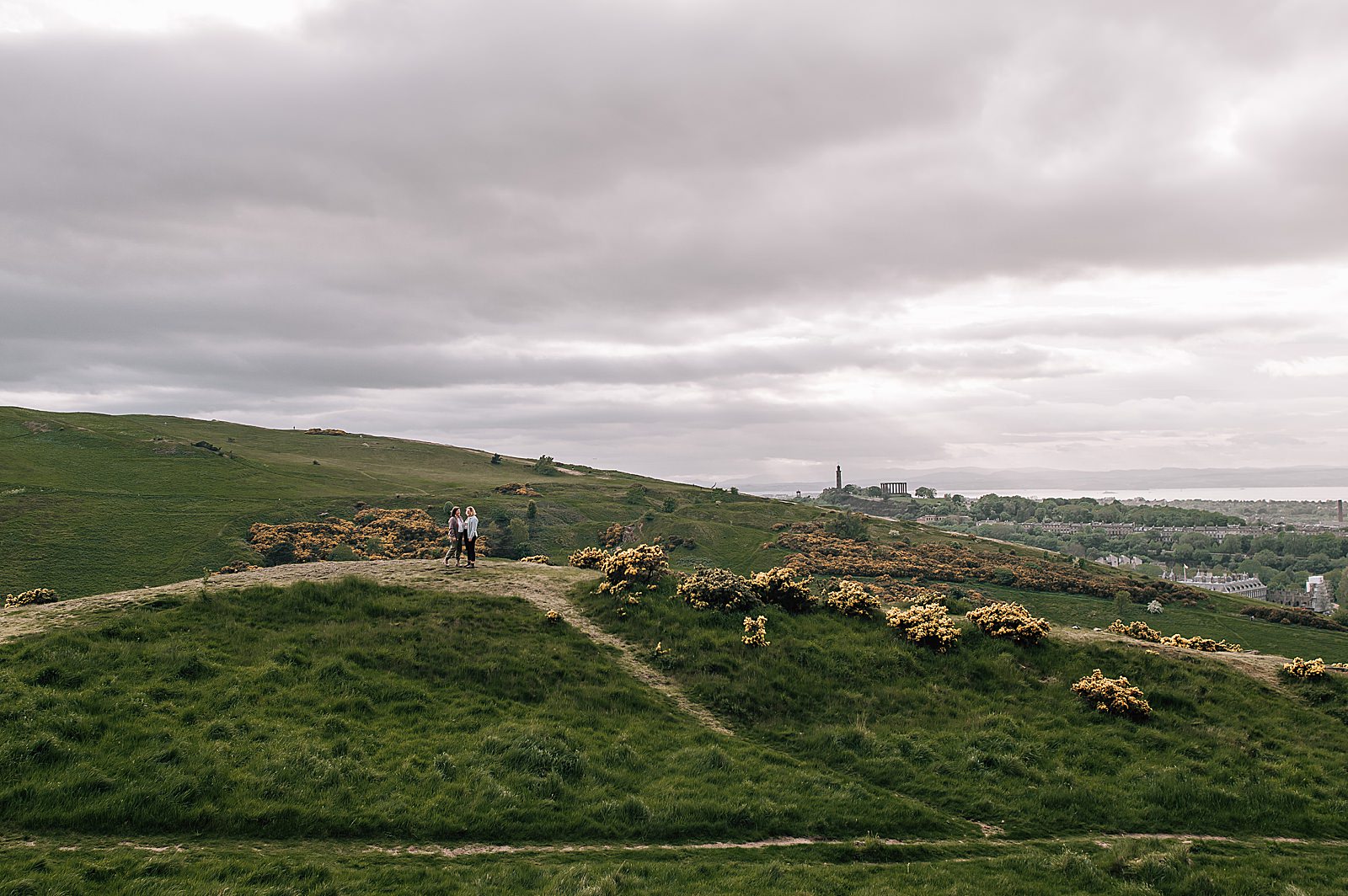 couple standing on green grassy hill with Edinburgh in the background captured by Edinburgh proposal photographer