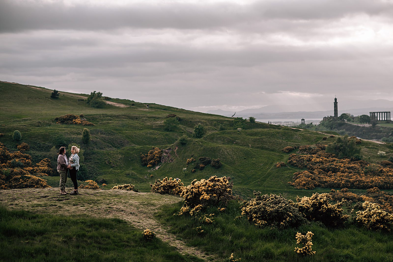 couple holding hands and looking at each other as they stand on top of a grassy hill with gorse bushes and Edinburgh in the back ground on a windy day captured by Edinburgh proposal photographer