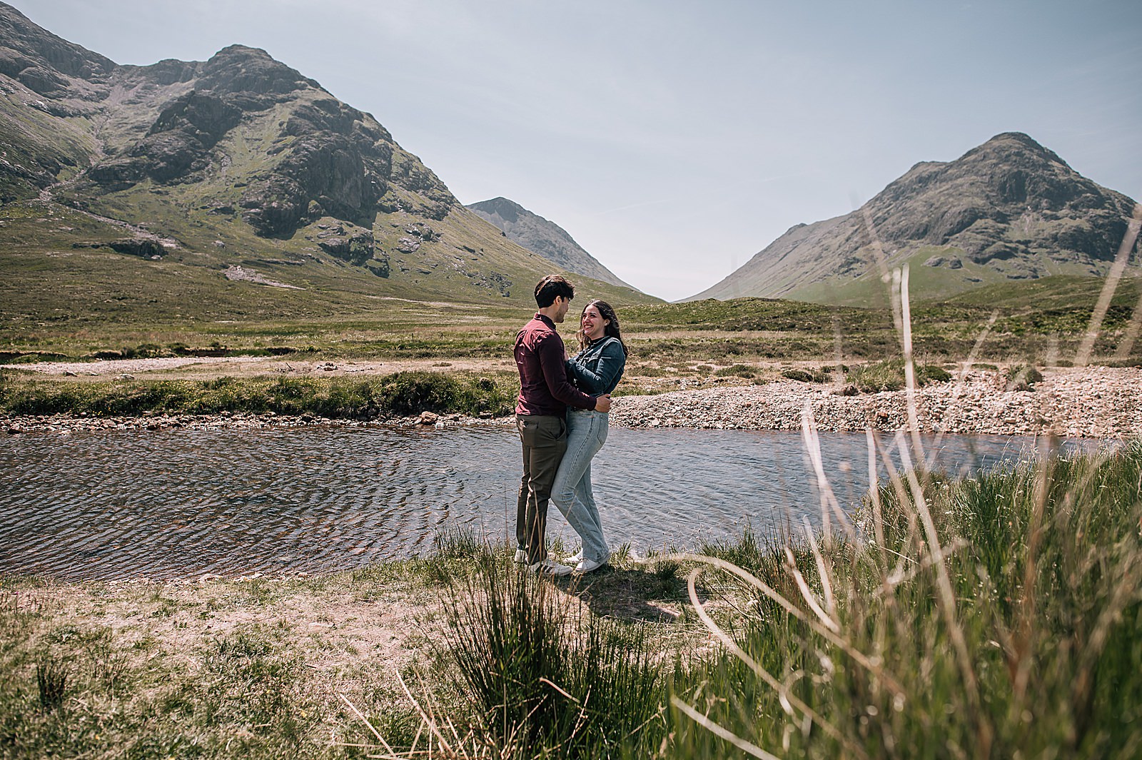 couple hugging and smiling by the side of a burn in Glencoe during their surprise proposal photographer photoshoot