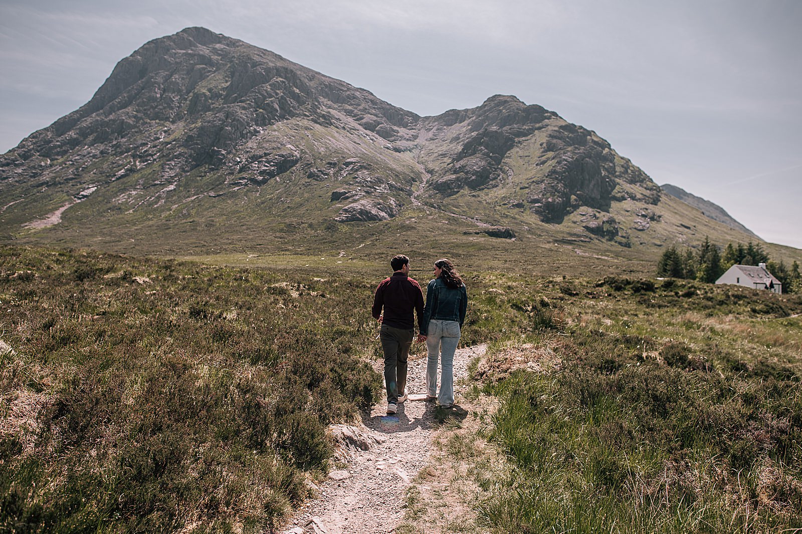 couple holding hands walking towards Lagangarbh hut in glencoe on a sunny day captured by Lagangarbh