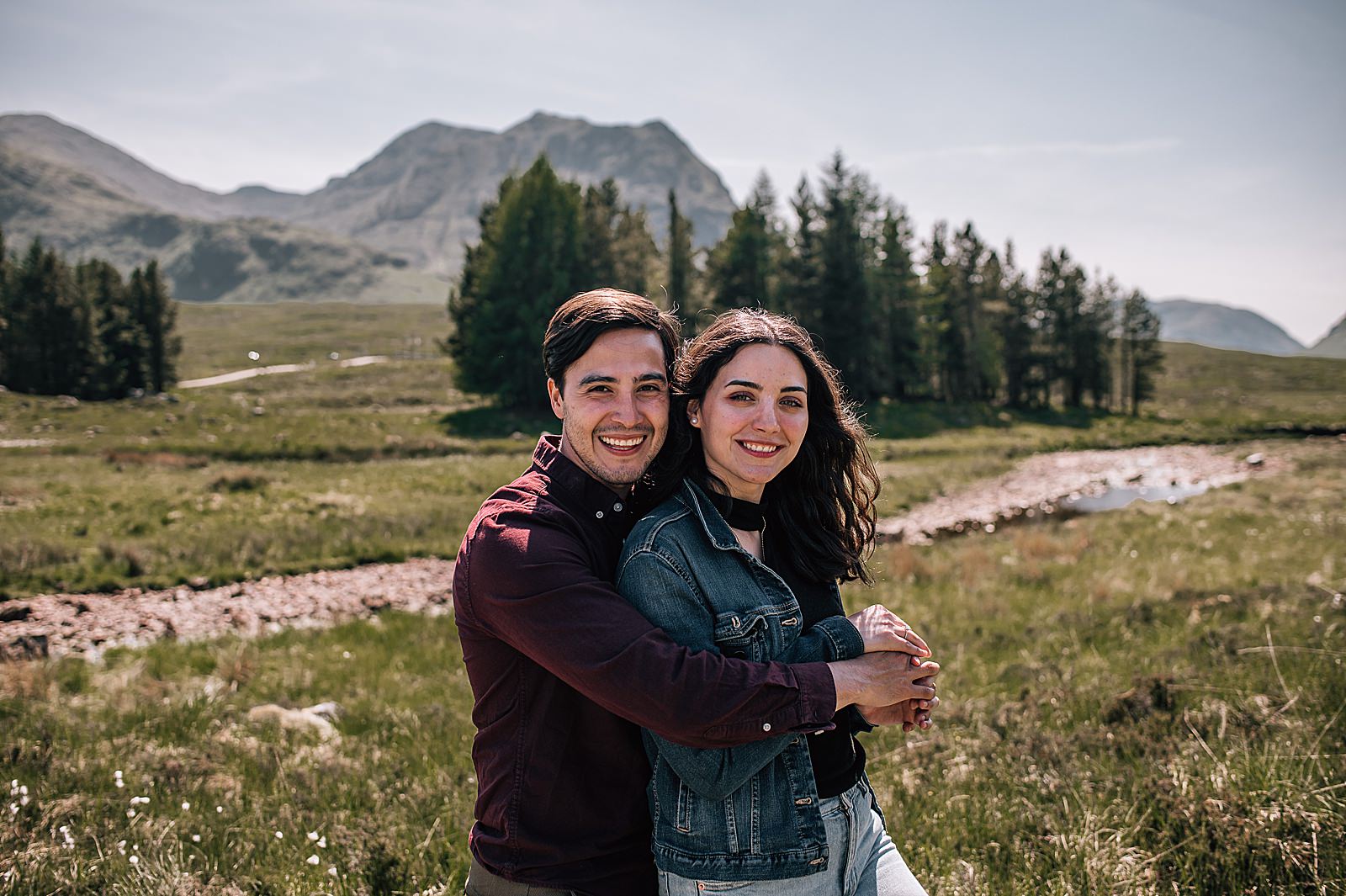 newly engaged couple smiling and hugging in front of stream and trees captured by glencoe proposal photographer