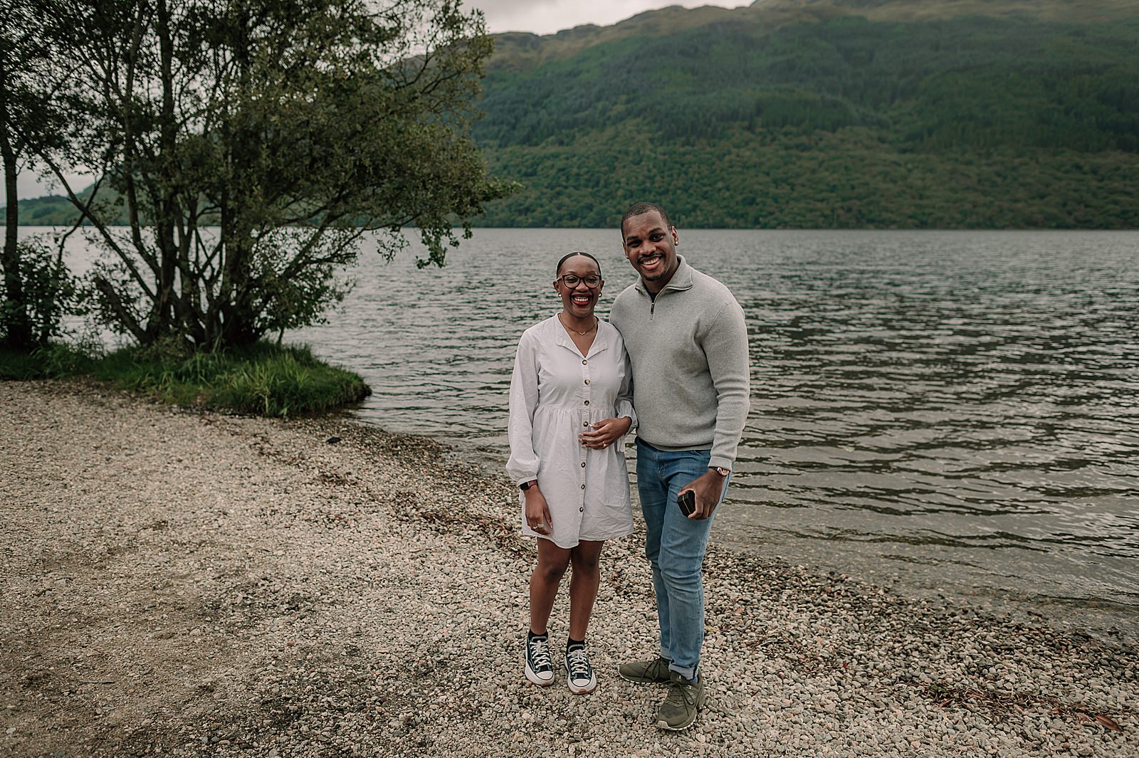 couple standing on the shore of Loch Lomond after getting engagement with surprise proposal captured by proposal photographer scotland
