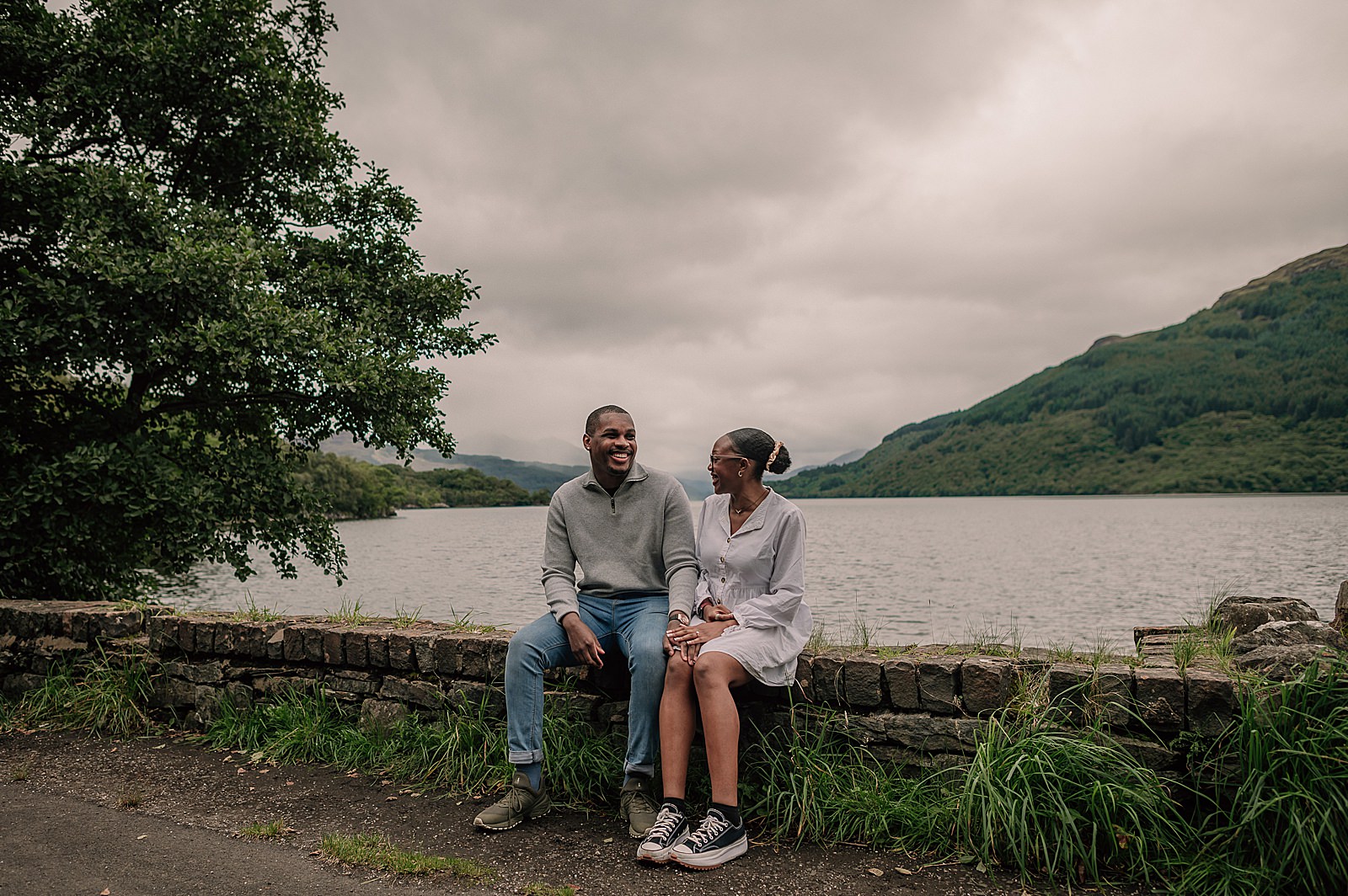 couple laughing sitting on the banks of loch lomond with green hills and grass after their engagement photography shoot