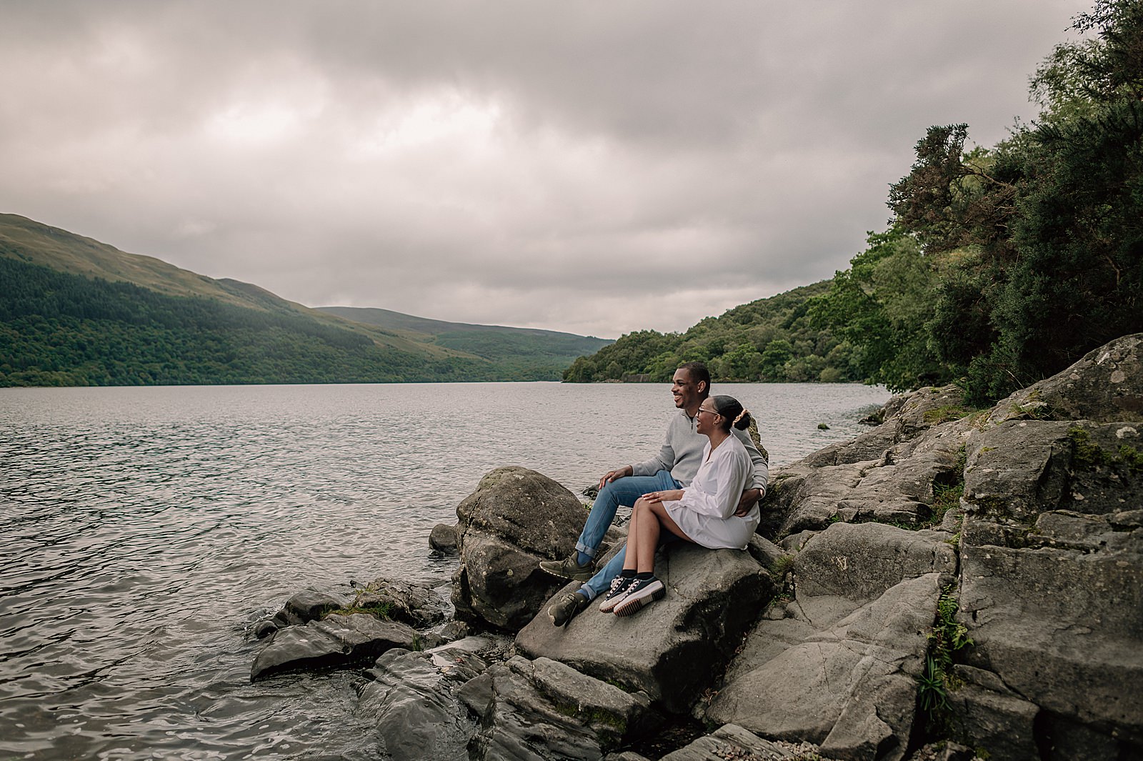 newly engaged couple sitting on rocks on the banks of Loch Lomond after surprise proposal captured by proposal photographer scotland