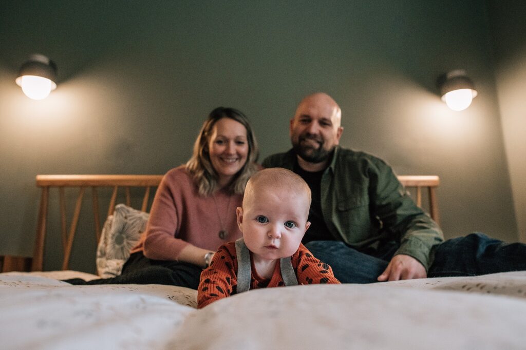 baby lying on tummy on a bed, lifting head and looking with mum and dad behind her