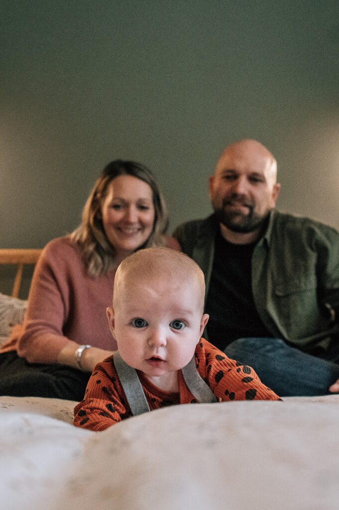 baby looking expressively at camera, lying on her tummy with mummy and daddy behind her