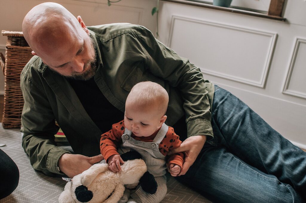 father lying behind baby as she's sitting up and playing with a toy