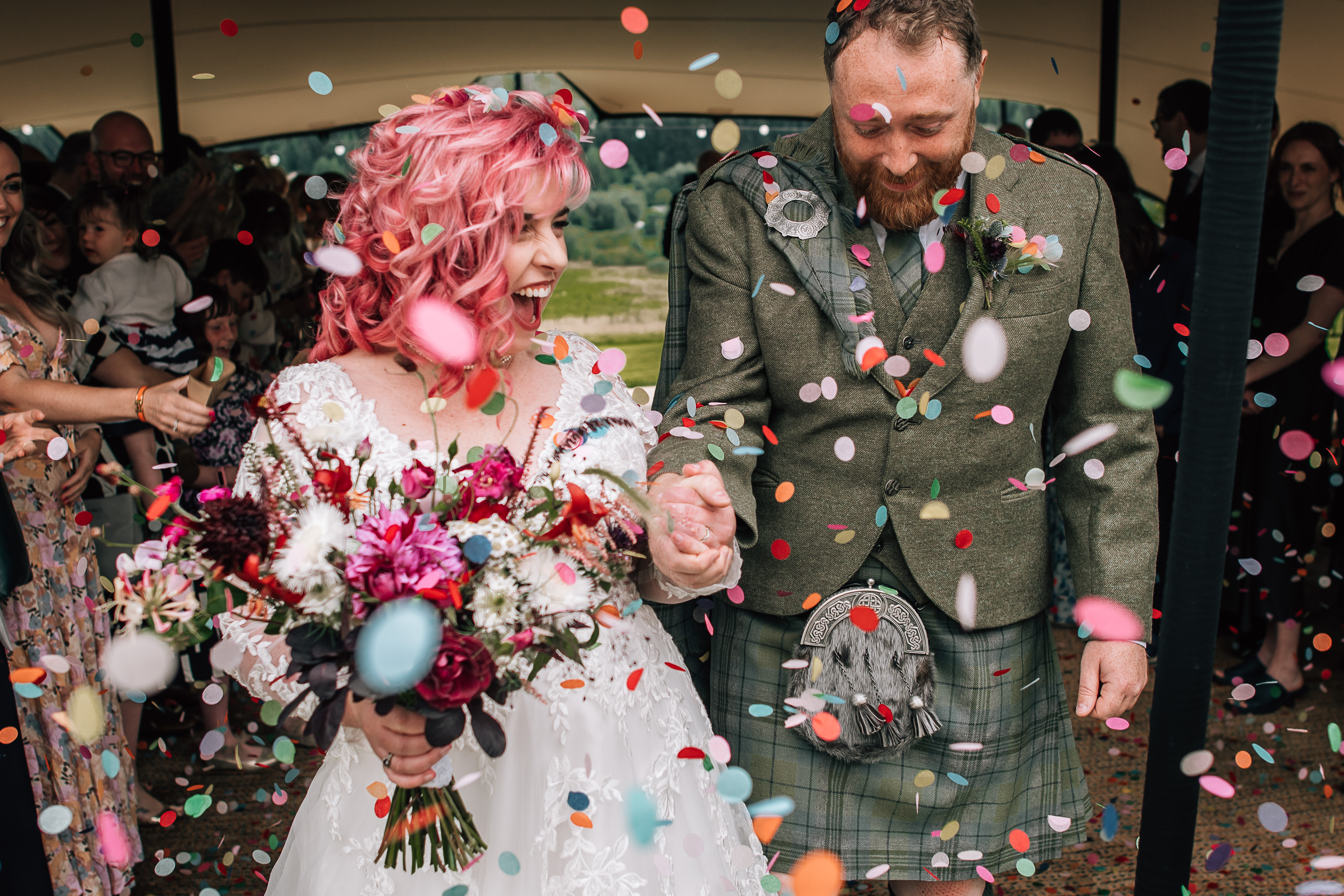 Couple laughing as they are surrounded by confetti- bride with pink hair and groom in kilt
