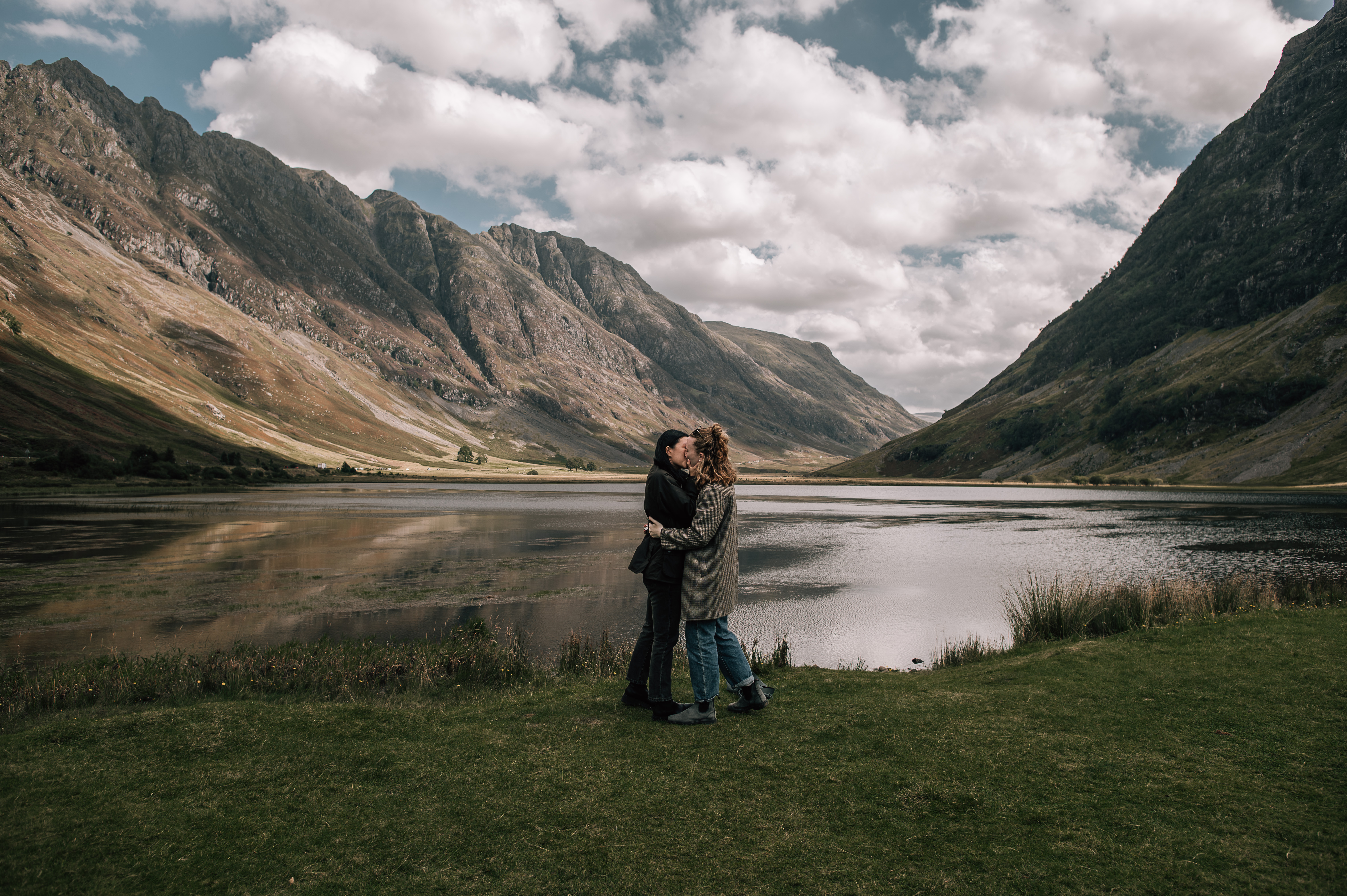 LGBTQ couple kissing in front of a lake, with mountains int he background, in Glencoe Scotland. 